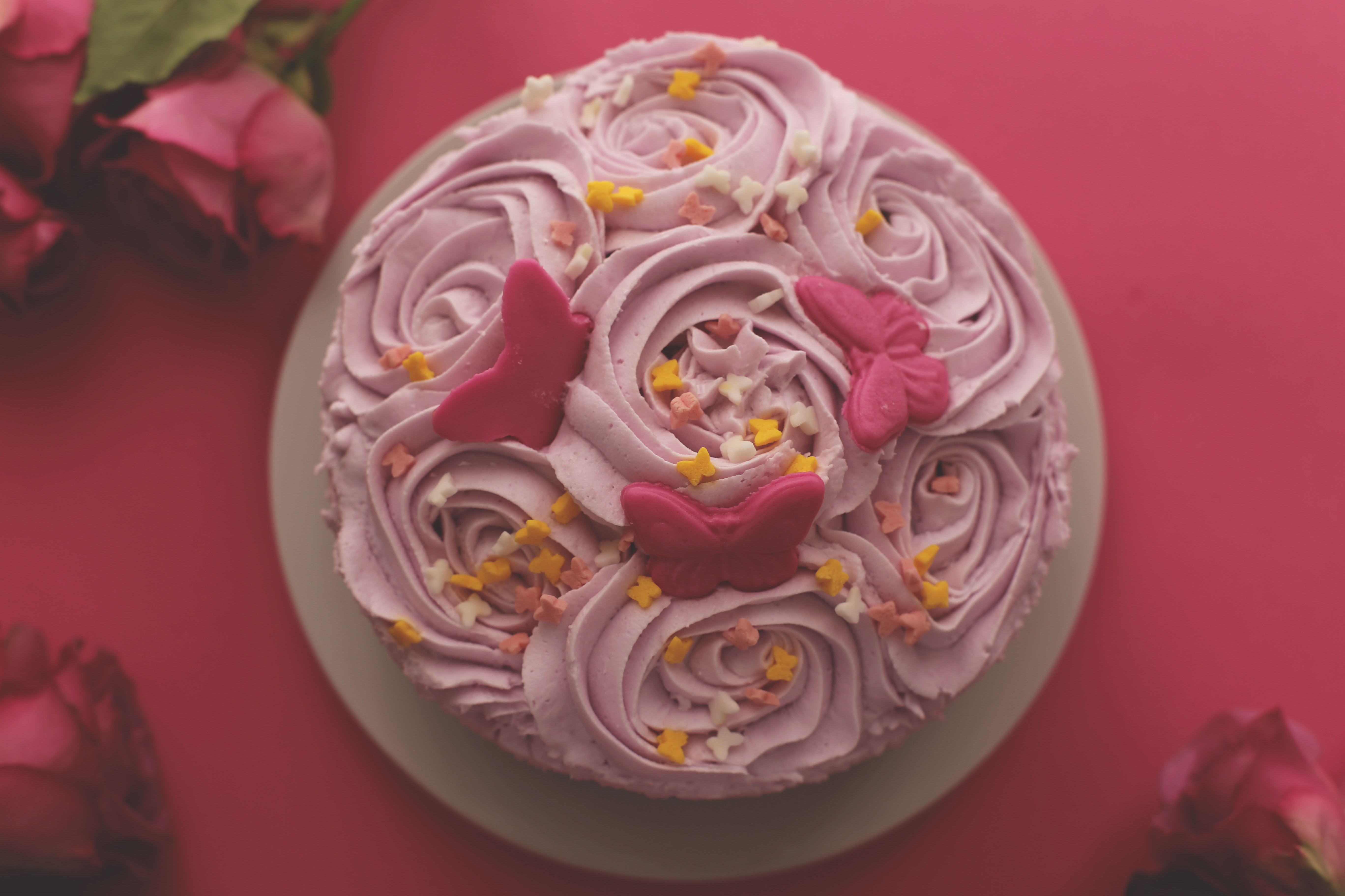 A pretty pink cake with swirl frosting, decorated with flowers and topped with adorable pink butterfly cookies on a red table.