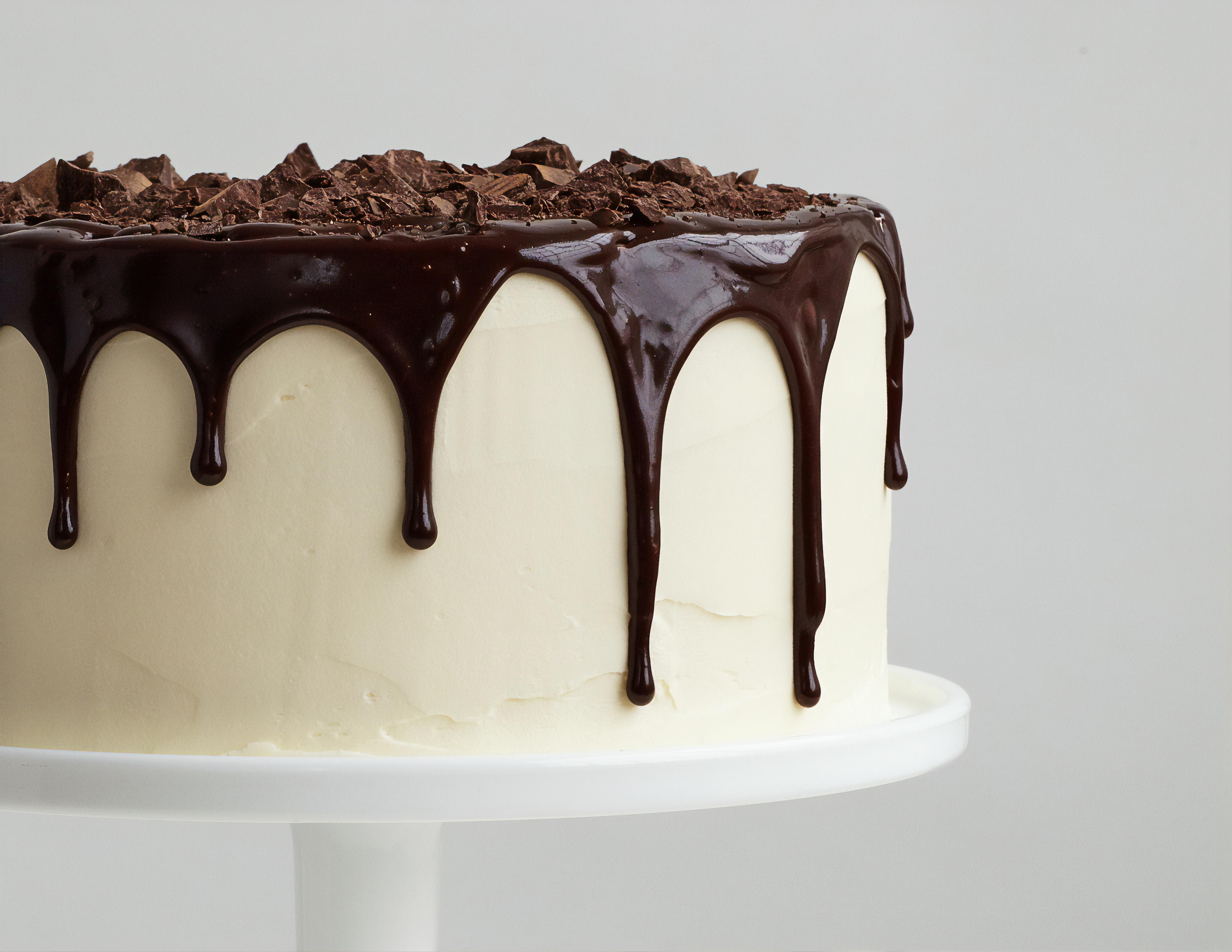 A delicious cake with smooth white sides and a luscious chocolate coating on top, dripping down the sides. It's adorned with chocolate shavings and sits on a pedestal.